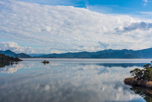 Trees with yellowed leaves on the shore of Koycegiz (Koycegiz) lake and reflections of clouds in the water. In the distance, a rocky and reedy islet can be seen.