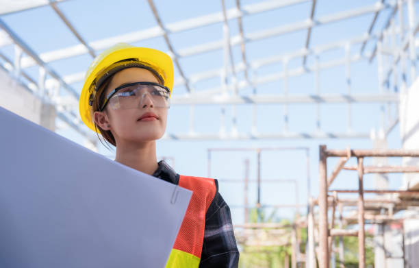 la ingeniera civil asiática usa un casco amarillo de seguridad con gafas y un plano de sujeción mientras revisa el edificio en el sitio de construcción. - architect women blueprint eyewear fotografías e imágenes de stock