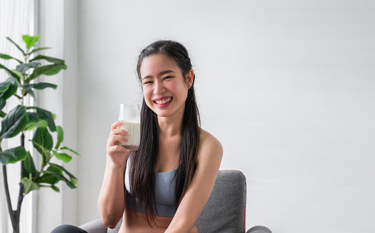 Happy Asian sports girl in sportswear sitting holding and drinking pure milk from glass after exercise in home. Healthy woman smiling and drink milkshake protein. Healthcare and weight loss concept.