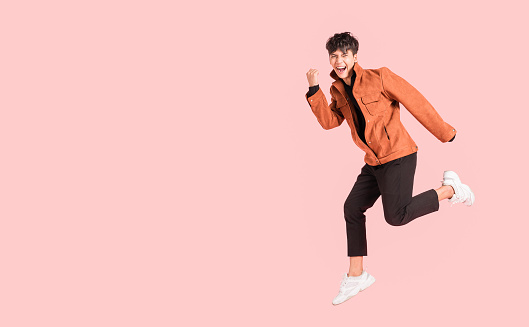 Happy handsome Asian man in fashionable clothing and jumping doing winner gesture isolated on pink background. Portrait of young male cheerful confident and excited jump in air and smile in studio.