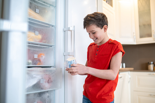 Smiling focused Caucasian teenager standing before an open freezer and examining a packaged food item