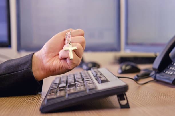 un hombre de negocios con una cruz religiosa católica en sus manos está trabajando en el teclado de una computadora en un escritorio de oficina, de cerca - desk job fotografías e imágenes de stock