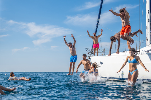 Group of young people jumping down from yacht into water. Enjoying party on yacht on sunny day. Summer vacation, youth and fun concept.