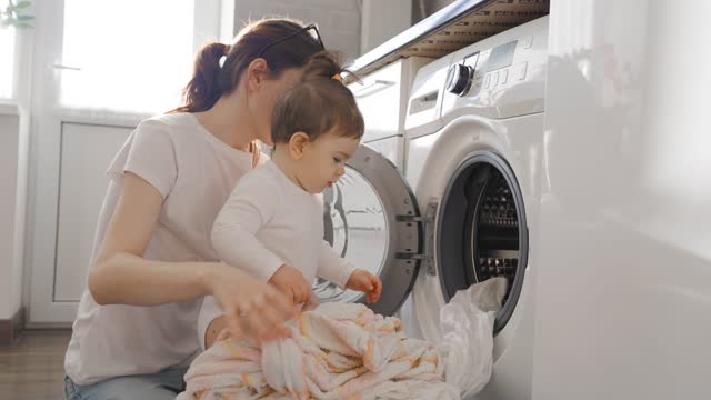 young mom take out clothes from washing machine with baby girl toddler in kitchen