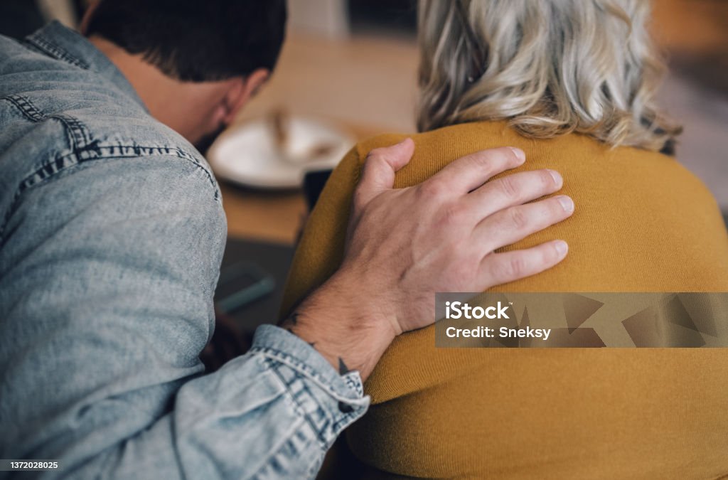 Son hugging his mother while sitting on the couch. Young man is lovingly holding an arm on his mother's shoulder while sitting together on their living room sofa. Shoulder Stock Photo