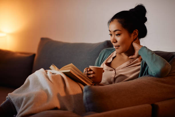 istock Young Asian woman dinking tea while reading a book on the sofa in the evening. 1372030854