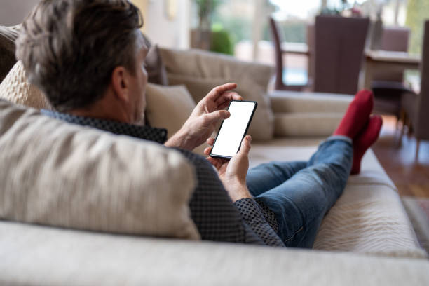 Caucasian man at home laying on the couch looking at smart phone Over the shoulder view of a caucasian man lying on a couch and looking at a smart phone with a white screen. man looking at phone over shoulder stock pictures, royalty-free photos & images