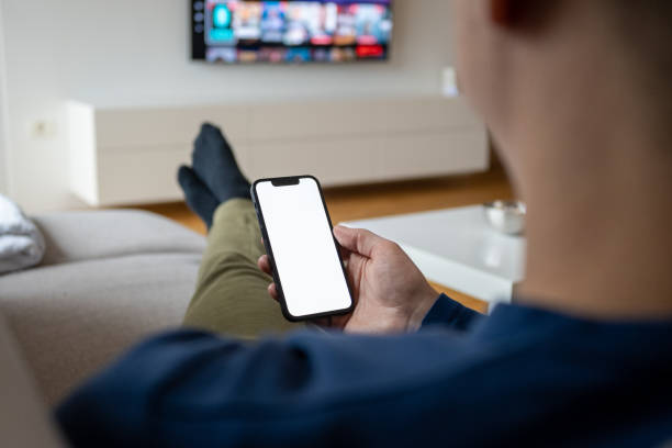 Caucasian man holding a smart phone while watching TV Over the shoulder view of a caucasian man holding a smart phone with a white screen while watching TV. legs crossed at ankle stock pictures, royalty-free photos & images