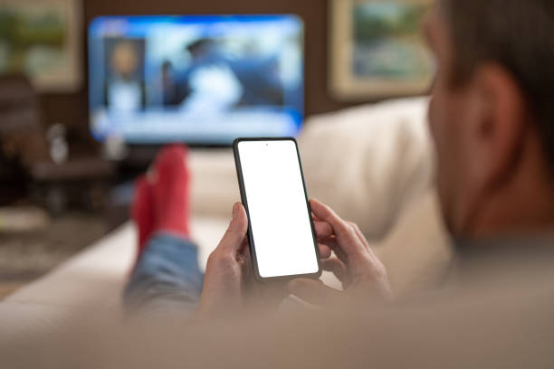 Caucasian man using a smart phone while watching TV Over the shoulder view of a caucasian man using a smart phone with a white screen while lying on a couch in the living room and watching TV. man looking at phone over shoulder stock pictures, royalty-free photos & images