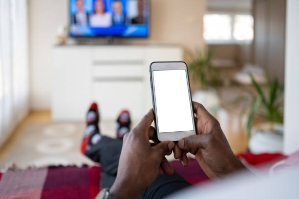 Black man using a smart phone while watching TV Black man using a smart phone with a white screen while lying with legs crossed on a sofa in the living room and watching TV. man looking at phone over shoulder stock pictures, royalty-free photos & images