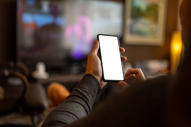 Caucasian man using a smart phone while watching TV Over the shoulder view of a caucasian man using a smart phone with a white screen while sitting on a couch in the living room and watching TV. man looking at phone over shoulder stock pictures, royalty-free photos & images