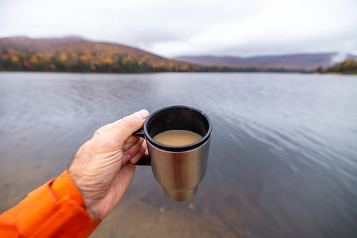 A man is drinking a coffee by the Monroe lake in camping in Autumn at Mont Tremblant National Park, Quebec, Canada. He is taking a break and contemplating the view. You can see his personal perspective of the nature surrounding him, the lake, the multi colored mountains with autumn leaf colors, etc…