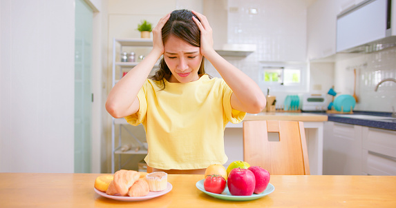 asian woman cannot decide to eat bread or fruits at home - Young girl chooses between fastfood and vegetable on the table