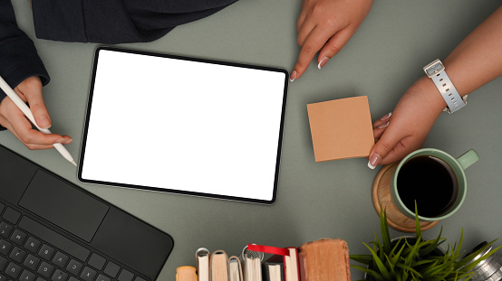 Two businesswomen discussing plan on digital tablet and working together in modern office desk. Tablet blank screen mockup, sticky notes, stationery and supplies. top view