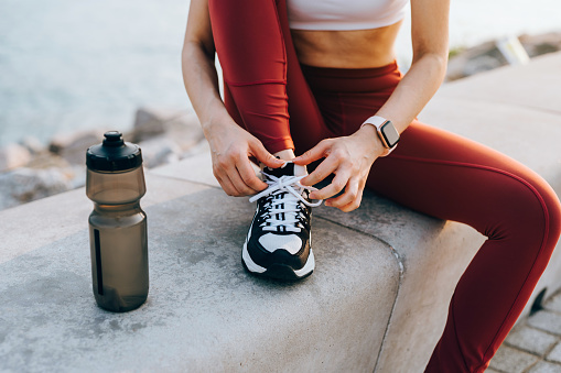 Cropped shot of active young Asian sportswoman in sports wear tying shoe laces and getting ready for a healthy jog outdoors in the city, with a bottle of water by her side. Active and healthy lifestyle. Outdoor workout. Health and fitness concept