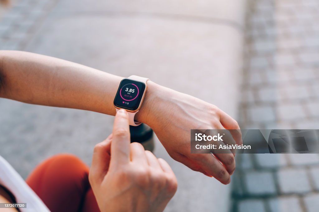 Over the shoulder view of young Asian sports woman checks her fitness statistics on smartwatch to monitor her training progress after working out in city outdoors. Active and healthy lifestyle. Outdoor workout. Health and fitness with technology concept Smart Watch Stock Photo