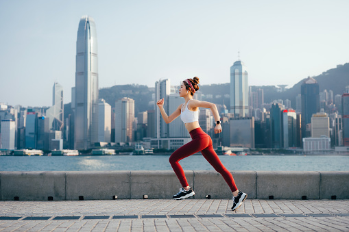 Side profile of active young Asian sports woman listening to music with headphones, running in urban park by the promenade in city at sunset. Active and healthy lifestyle. Outdoor running. Health and fitness concept