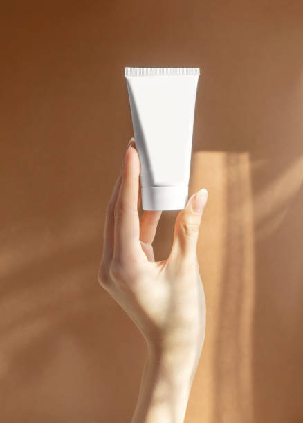 vertical image of woman's hand holding mock-up of white plastic cream tube in rays of sunlight. container for professional cosmetics. concept of skin care. close-up, brown background - moisturizer cosmetics merchandise human hand imagens e fotografias de stock