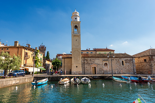 Lazise, Italy - May 26, 2021: Lazise village. Small port and ancient church of San Nicolo in Romanesque style. Tourist resort on the coast of Lake Garda, Verona province, Veneto, Italy, southern Europe. A small group of people strolls in front of the ancient monument on a sunny spring day.