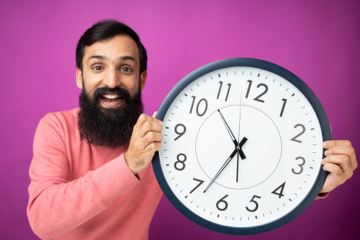 Picture of an excited man holding a clock near a purple wall smiling .