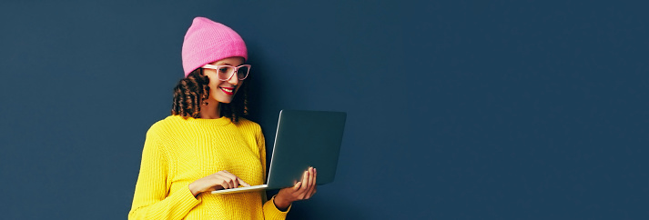 Portrait of stylish modern young woman working with laptop wearing colorful clothes on dark blue background, banner blank copy space for advertising text