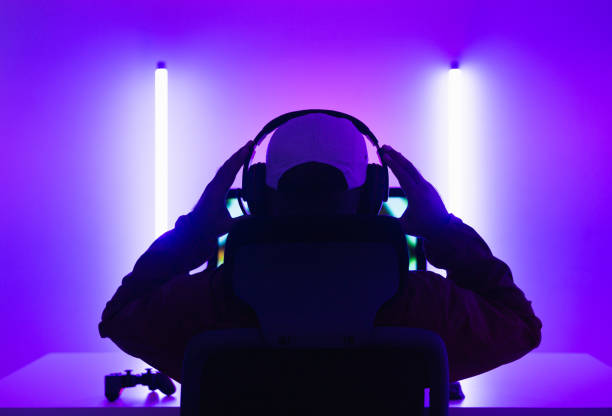 Rear view young Player video computer pc. Young man sitting on chair in game station.Streamer man wearing headphone  playing action game online in the darkroom.