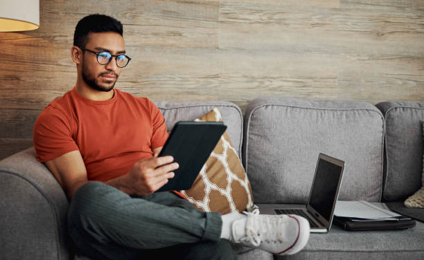 aufnahme eines gutaussehenden jungen mannes, der allein in seinem wohnzimmer sitzt und ein digitales tablet benutzt - people reading stock-fotos und bilder