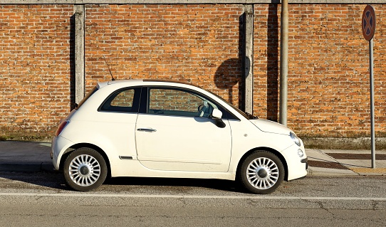 Udine, Italy. February 20, 2022. White Fiat 500 at the roadside. Brick wall and sidewalk on behind in front. Side view.