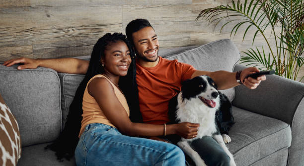 scatto di una giovane coppia felice seduta sul divano di casa con il loro border collie e guardando la televisione - couple young adult african descent multi ethnic group foto e immagini stock