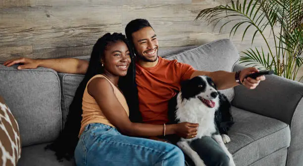 Photo of Shot of a happy young couple sitting on the sofa at home with their Border Collie and watching television