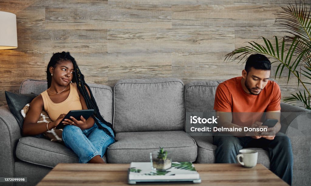 Shot of a handsome young man sitting and using technology in the living room while his boyfriend watches him He never gets off his phone! Infidelity Stock Photo