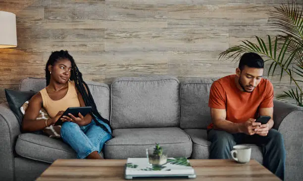 Photo of Shot of a handsome young man sitting and using technology in the living room while his boyfriend watches him