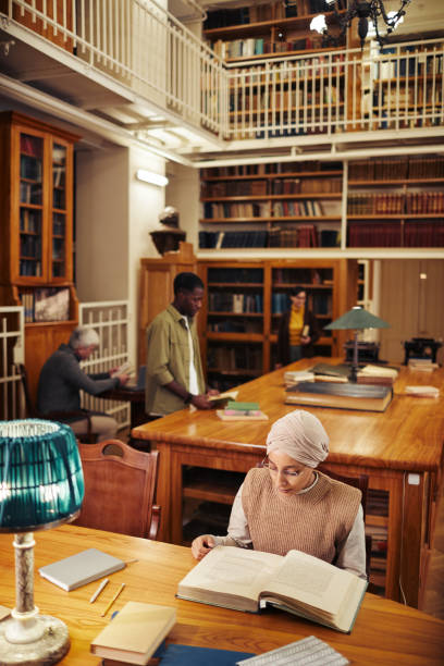 Group of People in Classic Library Vertical wide angle shot of classic college library with diverse group of people, focus on young woman wearing head covering and reading book middle eastern ethnicity mature adult book reading stock pictures, royalty-free photos & images