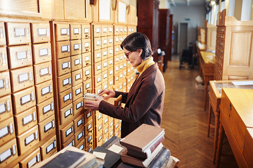 Side view portrait of adult woman searching for book in library catalogue cabinet, copy space