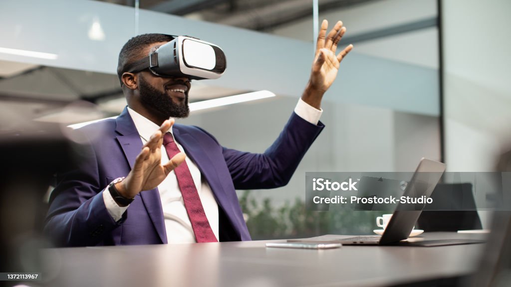 Excited black businessman sitting at workdesk, using VR glasses Modern technologies in business, virtual reality for enterprise concept. Excited black businessman sitting at workdesk in front of laptop, using VR glasses, touching copy space, modern office interior Virtual Reality Stock Photo