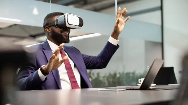 Modern technologies in business, virtual reality for enterprise concept. Excited black businessman sitting at workdesk in front of laptop, using VR glasses, touching copy space, modern office interior