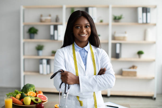 Portrait of friendly african american nutritionist with measuring tape and stethoscope recommending healthy eating Portrait of friendly african american nutritionist with measuring tape and stethoscope in hand recommending healthy eating and diet, posing in clinic office nutritionist stock pictures, royalty-free photos & images