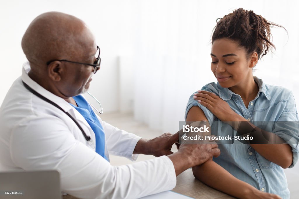 Pregnant Black Lady Getting Vaccinated, Doctor Applying Adhesive Bandage Antiviral Vaccination, Immunization. Mature medical worker sticking plaster bandage on patient's shoulder after injection in health centre. Pregnant black woman getting vaccine for Covid prevention Vaccination Stock Photo