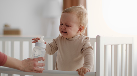 Lindo bebé infeliz llorando en la cuna, la mano de la madre dando biberón con agua o leche al niño pequeño photo