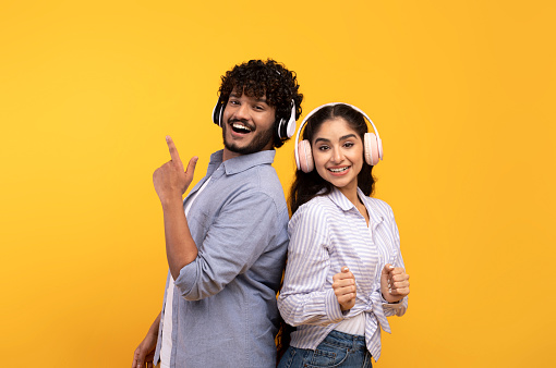 Enjoy music together. Happy young indian couple in wireless headphones listening songs and dancing, smiling at camera on yellow background, studio shot
