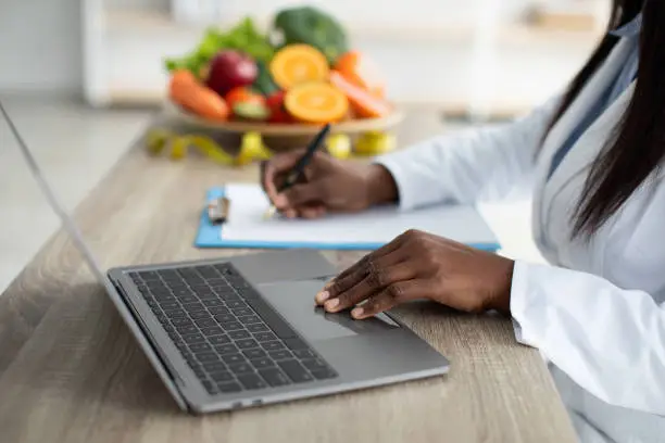 Working process. African american dietitian creating individual diet plan for patient or counting calories, working on laptop and clipboard, selective focus, closeup