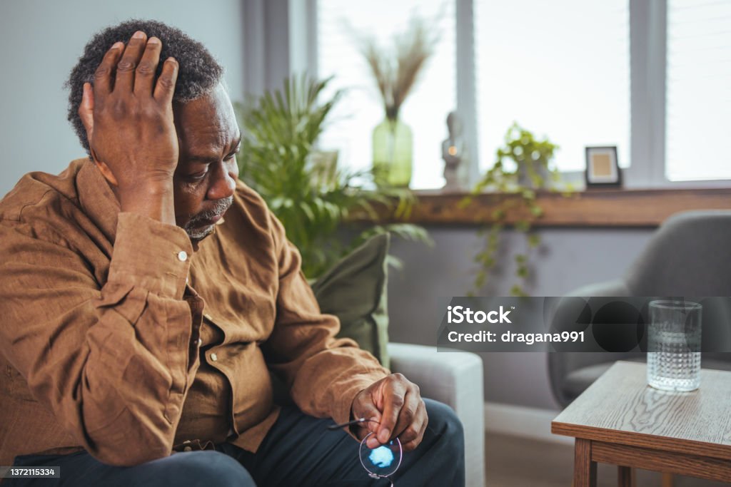 Unhappy middle aged retired grandfather suffering from headache disease indoors. Frustrated unhealthy senior mature man touching head, having painful feelings sitting alone at home. Unhappy middle aged retired grandfather suffering from headache disease indoors. Loneliness Stock Photo