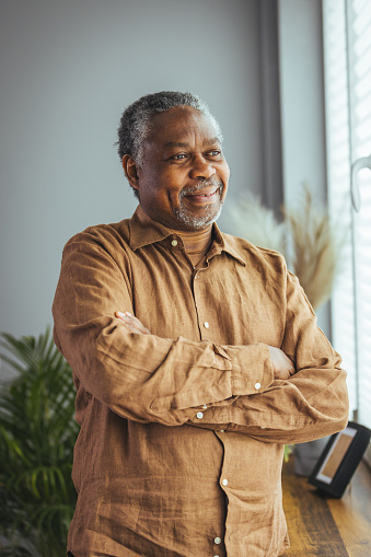 Side view of a happy African retired elderly man standing at home by the window. A satisfied old man looks out the window and smiles as he stands by the window. Positive and self-confident seniors enjoy retirement.