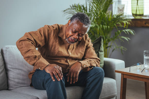 Mature man massaging his painful knee. Photo of mature, elderly man sitting on a sofa in the living room at home and touching his knee by the pain during the day. Mature man massaging his painful knee. knee stock pictures, royalty-free photos & images