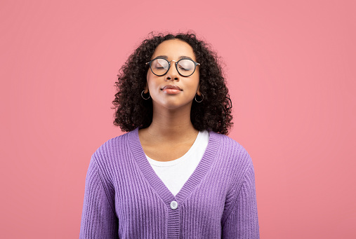 Calm young black woman in glasses closing her eyes, breathing freely on pink studio background. Peaceful millennial African American lady meditating, feeling relaxed and balanced