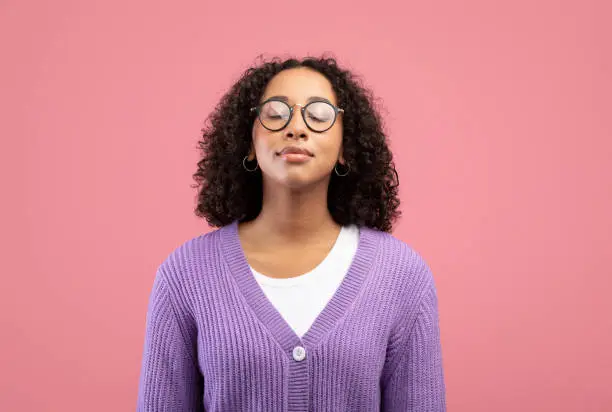 Photo of Calm young black woman in glasses closing her eyes, breathing freely on pink studio background