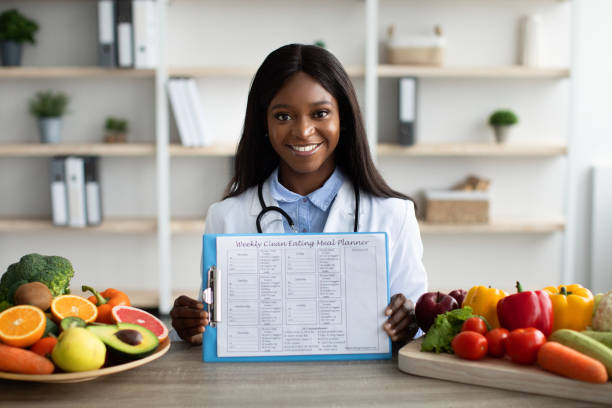 istock Dieting concept. Young dietitian showing weekly diet plan, sitting at desk with plates of fruits and vegetables 1372116329
