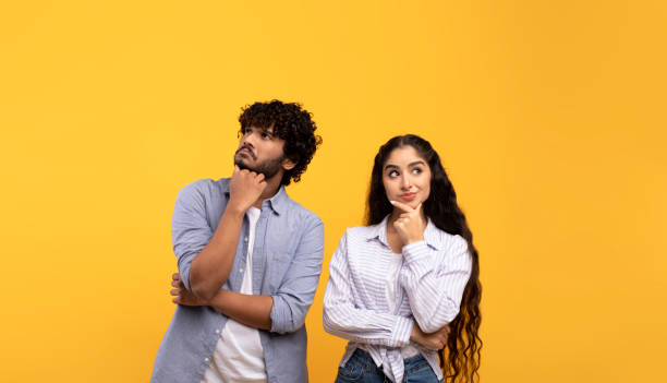 Portrait of pensive indian man and woman thinking and looking up at free space, touching chin, yellow background stock photo
