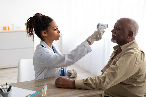 Black Medical Worker Doing Temperature Screening For Elderly Patient Man Measuring Fever Before Vaccination Using Contactless Infrared Themometer. Coronavirus Protection In Hospital Concept