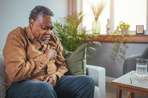 African senior man sitting on sofa at home with sickness. Elderly male suffering from heart attack and feel unwell need for medicine. Retirement people health care and illness recovery concept
